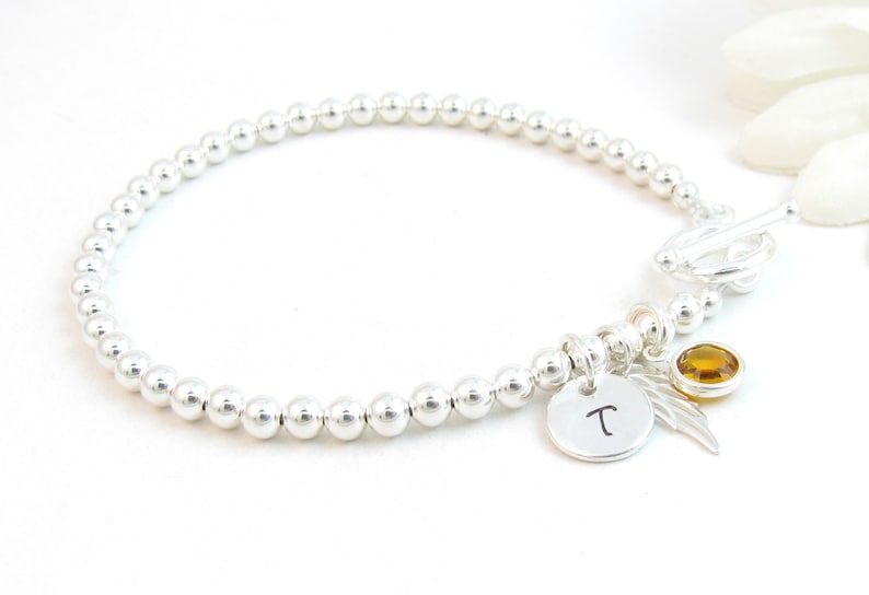 May include: Silver bead bracelet with a charm that has the letter "T" on it, a small amber gemstone, and a silver angel wing charm.