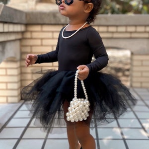 May include: A young girl wearing a black tulle tutu, black long-sleeved top, and black shoes. She is wearing a pearl necklace, a pearl handbag, and oversized black sunglasses. She is also wearing a silver tiara.