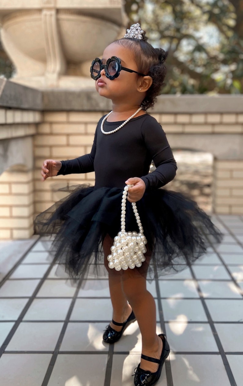 May include: A young girl wearing a black tulle tutu, black long-sleeved top, and black shoes. She is wearing a pearl necklace, a pearl handbag, and oversized black sunglasses. She is also wearing a silver tiara.