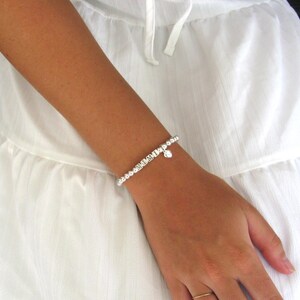 May include: A silver beaded bracelet with a small heart charm is worn on a wrist. The bracelet is made of small, round beads and is paired with a white dress. The bracelet is a delicate piece of jewelry.