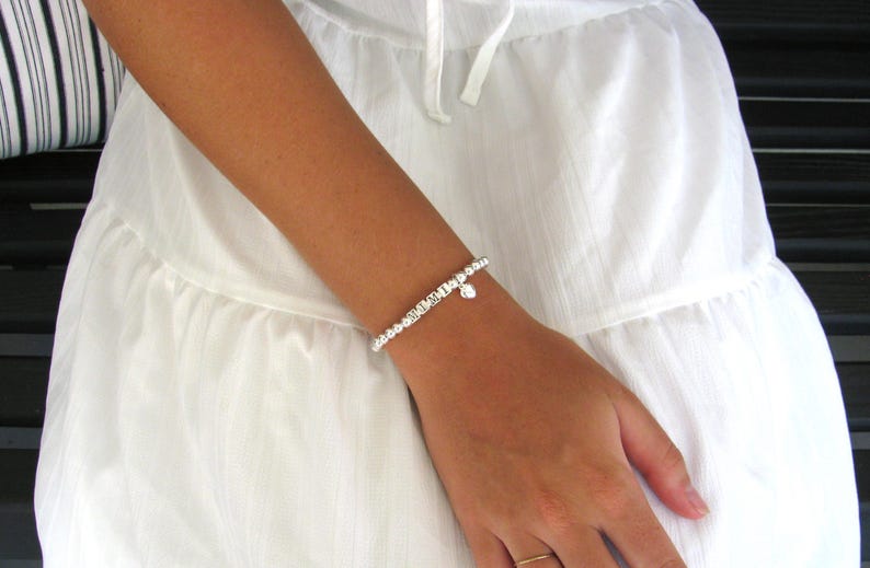 May include: A silver beaded bracelet with a small heart charm is worn on a wrist. The bracelet is made of small, round beads and is paired with a white dress. The bracelet is a delicate piece of jewelry.