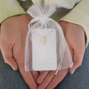 May include: A delicate gold necklace featuring a small letter charm and a feather pendant, presented on a white card with the text "pickled beans handmade jewellery". The necklace is packaged in a white organza bag.
