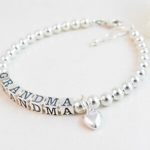May include: A silver beaded bracelet with the word "GRANDMA" spelled out in square letter beads. A small heart charm dangles from the bracelet. The bracelet is on a white surface.