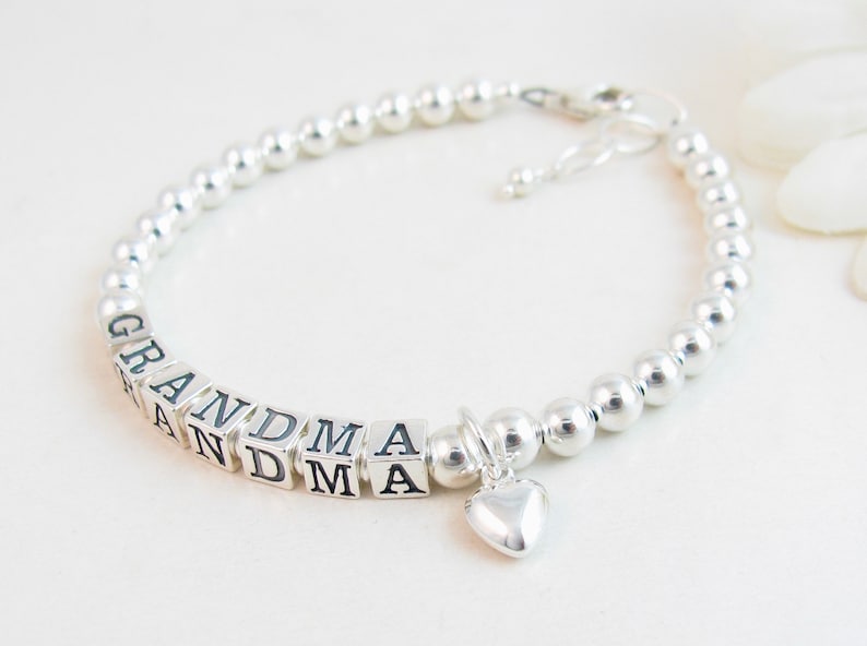 May include: A silver beaded bracelet with the word "GRANDMA" spelled out in square letter beads. A small heart charm dangles from the bracelet. The bracelet is on a white surface.