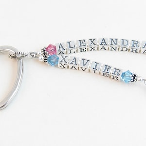 May include: A silver heart-shaped keychain with two strands of beads spelling out the names "ALEXANDRA" and "XAVIER". The beads are white with black lettering and are accented with pink and blue crystals.