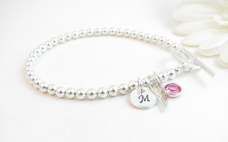 May include: A silver beaded bracelet with a toggle clasp. The bracelet has a charm with the letter "M" and a small pink crystal.
