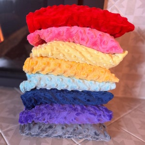May include: A stack of seven plush, textured pillows in a variety of colours: red, pink, yellow, orange, light blue, navy blue, purple, and grey. The pillows are stacked on top of each other. A fireplace is in the background.