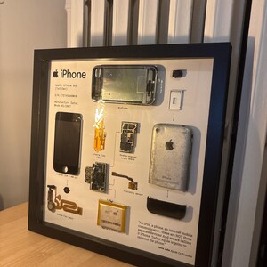 以下が含まれることがあります： 黒いフレームに収められた分解済みの初代Apple iPhone。ロジックボード、バッテリー、筐体などの内部コンポーネントが白い背景に配置され、説明文とAppleロゴが添えられています。