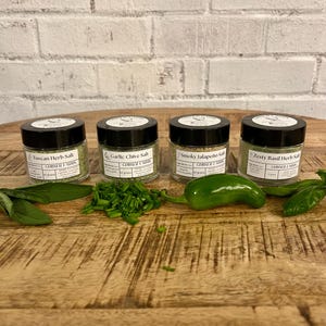 May include: Four small glass jars with black lids, labeled Tuscan Herb Salt, Garlic Chive Salt, Smoky Jalapeno Salt, and Zesty Basil Herb Salt. Fresh herbs and a green pepper are arranged in front of the jars on a wooden surface.