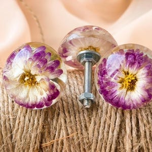 以下が含まれることがあります： 紫と白の花が中に閉じ込められた、透明な樹脂製のキャビネットノブが3つ。ノブは金属製のボルトに取り付けられ、茶色の紐のコイルの上に置かれています。花の中心は黄色です。