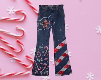 May include: Blue denim jeans with a festive Christmas design. The jeans feature a gingerbread man, candy canes, and snowflake accents. The flared legs have red and white striped patterns, creating a holiday-themed look.