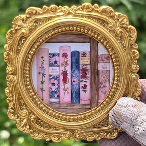 May include: A circular print of a bookshelf featuring six book spines with floral illustrations, housed in an ornate, metallic gold frame with intricate scrollwork and beaded detailing. The books display various pink, blue, and red botanical designs.