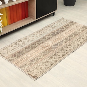 May include: Rectangular beige rug with a geometric pattern of stripes and zigzags. The rug has a distressed look with faded areas, and is placed on a light wood floor. A wooden shelf with books and decorative items is in the background.