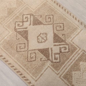 May include: Beige and brown patterned rug with geometric designs. The rug features a central square design with a smaller square in the center, surrounded by a border of repeating patterns. The rug has a distressed, vintage look.