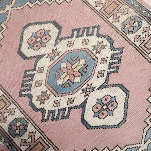 May include: A rectangular, vintage-style rug with a central pink field and intricate geometric patterns. The design incorporates light blue, beige, and dark blue accents, creating a symmetrical and detailed composition. The rug's edges feature a border with repeating motifs.