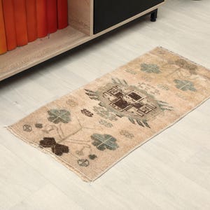 May include: A rectangular area rug featuring a beige base color with a central dark brown geometric medallion and symmetrical floral motifs in muted green and brown tones. The rug has short fringe on the ends and a low-pile woven texture.