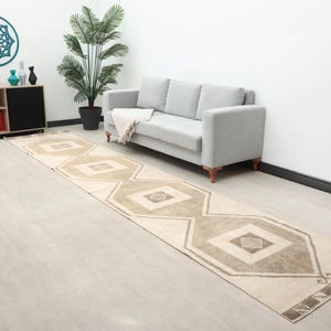 Op de afbeelding: Een lange, rechthoekige vloerkleed met een geometrisch patroon in beige en bruine tinten. Het vloerkleed heeft herhalende ruit- en rechthoekige vormen. Het vloerkleed ligt op een lichtgekleurde houten vloer, met een bank en decoratieve items op de achtergrond.