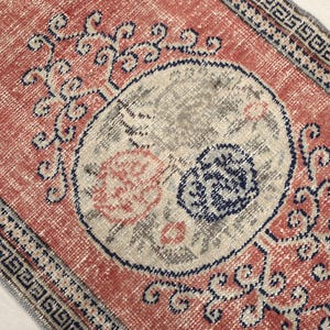 May include: A vintage rug with a red background and a large, circular, off-white design featuring a stylized floral pattern. The rug has a blue and white border with a geometric pattern.