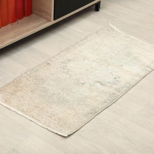 May include: A rectangular, off-white rug with a faded, mottled pattern. The rug has a slightly distressed appearance and a fringed edge. The rug is placed on a light-coloured wooden floor, near a shelf with books.