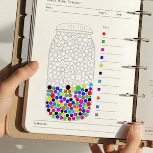 Può includere: Un'agenda beige aperta su una pagina con un design "Small Wins Tracker". Il tracker presenta un'illustrazione di un barattolo pieno di cerchi colorati, con una chiave di quadrati colorati. La parte superiore della pagina ha spazi per "Goal", "Start" e "End".