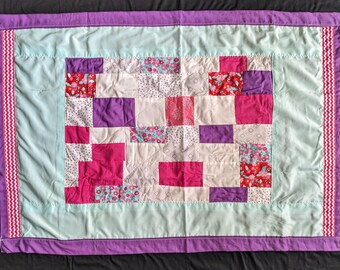 May include: A patchwork quilt with a light blue background and a variety of pink, purple, and white squares. The quilt is edged with a purple border and a pink and white border.