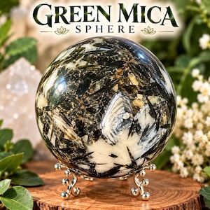Op de afbeelding: Een gepolijste groene mica bol met donkere, naaldachtige insluitingen en witte mineraalvlekken. De bol rust op een zilverkleurige metalen standaard. De tekst "GREEN MICA SPHERE" staat in donkergroene letters boven het object.