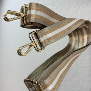 May include: A replacement bag strap featuring a woven pattern of beige, metallic gold, and white stripes. The strap is equipped with polished gold-tone metal lobster claw clasps at both ends, allowing for easy attachment to various handbags or accessories.