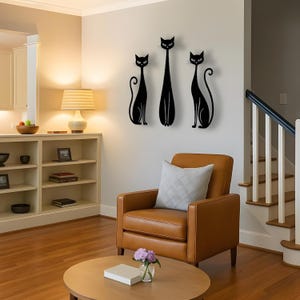 May include: A set of three black wall-mounted cat silhouettes featuring elongated bodies and curled tails. The figures are arranged vertically on a light-colored wall, displaying stylized feline forms with pointed ears and almond-shaped eyes.