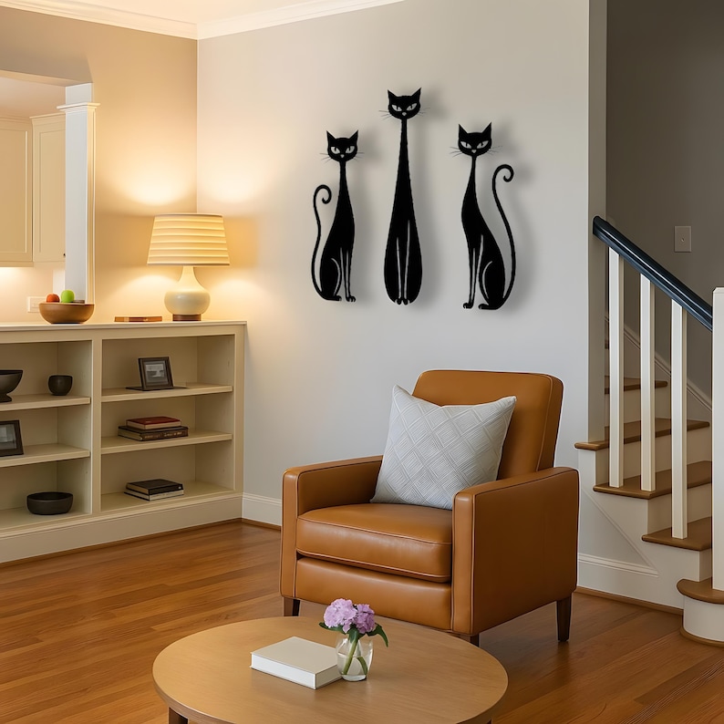 May include: A set of three black wall-mounted cat silhouettes featuring elongated bodies and curled tails. The figures are arranged vertically on a light-colored wall, displaying stylized feline forms with pointed ears and almond-shaped eyes.