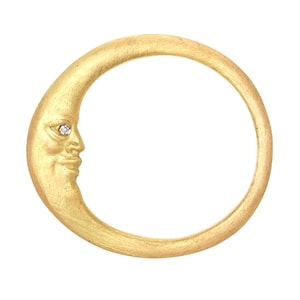 May include: A gold-colored crescent moon ring with a face carved into the moon's surface. A small, sparkling gemstone is set in the eye of the moon. The ring is a celestial-themed piece of jewelry.