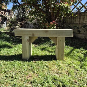 May include: A light-coloured wooden bench with a rectangular seat and two supporting legs. The bench is constructed from unfinished timber and is set on a grassy lawn. The bench is approximately 122 cm long.