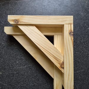 May include: A wooden corner bracket made from light-coloured pine. The bracket has a right-angle design with a diagonal support beam. The wood grain and knots are visible, showing the natural material. Suitable for shelving or other support needs.