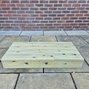May include: A rectangular wooden step made from light-coloured planks. The step is constructed from multiple parallel wooden boards, secured together. It is placed on a grey stone surface, with a red brick wall in the background. The step is approximately 75 cm long.