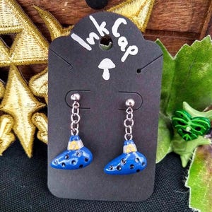 May include: A pair of blue ocarina earrings with silver chains and hooks. The ocarinas have a yellow and white design on the top and are displayed on a black card with the text "Ink Cap" written on it.