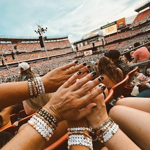 May include: Several arms are raised, displaying multiple bracelets with white and gold beads. Some bracelets spell out names like "MORGAN WALLEN." The background shows a crowded stadium with a concert stage.