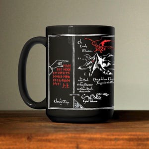 May include: Black ceramic mug featuring a map design with white and red illustrations and text. The map includes a mountain, a dragon, and text that reads "Thror's Map". The mug is on a wooden surface.