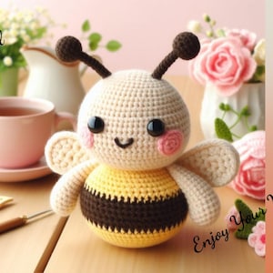 May include: A crocheted bee toy with a yellow and brown striped body, cream colored wings, and a cute face with black eyes and pink cheeks. The bee is sitting on a wooden surface with a cup of tea and flowers in the background.