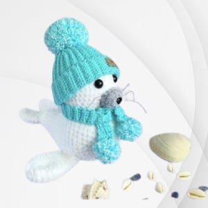 May include: A white crocheted seal wearing a blue knitted hat and scarf with a pom-pom on top. The seal is sitting on a white background with seashells scattered around it. The text "Sweet Seal" is written in blue above the seal and "BKT Digital" is written in blue below the seal.