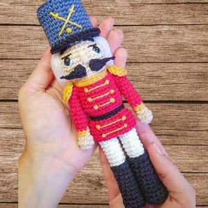May include: A crocheted toy soldier with a red and gold uniform, a blue hat with crossed swords, and a white face with black eyes, mustache, and mouth. The toy is held in two hands against a wooden background. The text "Tin Soldier" is above the toy and "BKT Digital" is below the toy.