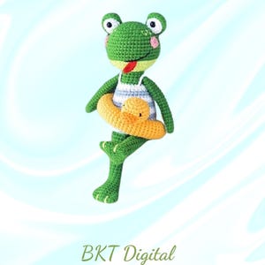 May include: A green crocheted frog wearing a blue and white striped swimsuit and holding a yellow crocheted life preserver. The frog is sitting on a blue and white background with the text "Swimming Frog" and "BKT Digital" in green.