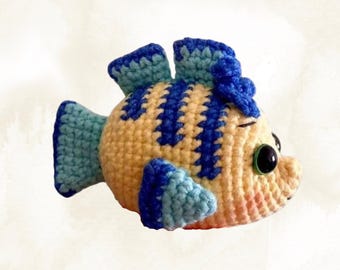 Puede incluir: Un juguete de pez de aleta azul tejido a crochet con hilo amarillo, azul y azul claro. El pez tiene ojos negros grandes y un pequeño lazo azul en la cabeza. El texto "Blue-Finned Fish" está por encima del pez y "BKT Digital" está por debajo del pez.