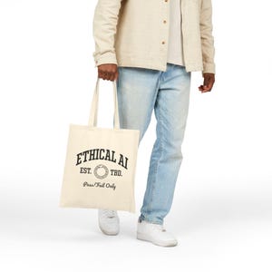 May include: A cream-coloured tote bag featuring the text "ETHICAL AI" in black. Below, it reads "EST. TBD." and "Pass/Fail Only." The bag is carried by a person wearing light blue jeans and a beige jacket.