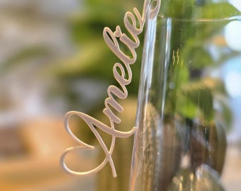 May include: A clear wine glass with a decorative name tag that reads "Amelie" in cursive script. The tag is attached to the rim of the glass. A lit candle and greenery are visible in the background.
