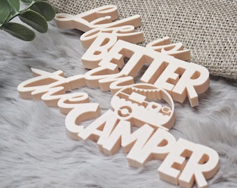 May include: A light beige wooden sign with the words "Life is Better in the Camper". The sign has a camper graphic and is placed on a gray furry surface with a burlap background and green leaves.