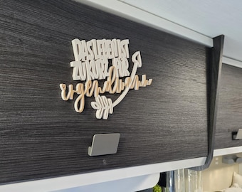 May include: A wooden sign with the German phrase "DAS LEBEN IST ZUKURZ FÜR IRGENDWANN" in white and light brown. The sign is mounted on a dark gray cabinet door. The phrase translates to "Life is too short for someday."