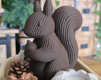 May include: A dark brown, 3D-printed squirrel figurine holding an acorn, featuring a distinct ribbed texture throughout its body. The squirrel is positioned next to two brown pinecones and a white, ribbed, circular candle holder on a tray.