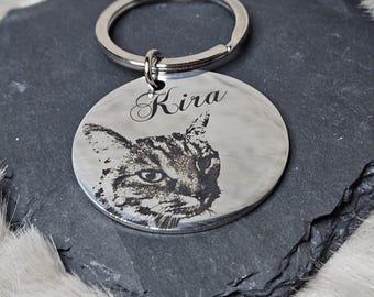 May include: A round, silver-colored keychain tag with a detailed engraved image of a cat's face. The name "Kira" is elegantly scripted above the cat's image. The keychain has a silver ring for attachment.