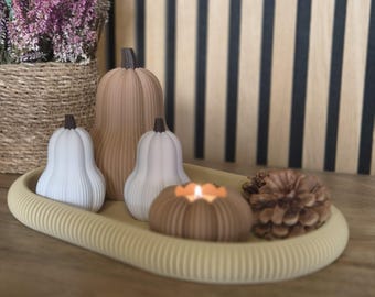 May include: A beige oval ribbed tray holding two white pear-shaped pumpkins, one tall tan ribbed pumpkin, a small brown pumpkin-shaped candle with a lit flame, and a brown pinecone. The items are arranged together on the textured surface.