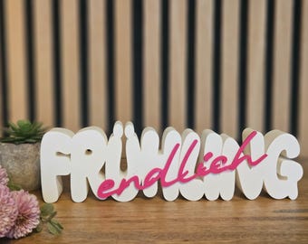 May include: A white wooden sign with the word "FRÜHLING" in large, rounded letters, with "endlich" in pink script overlaying it. The sign is on a wooden surface, with a small potted succulent and pink flowers in the foreground.
