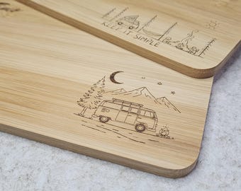 May include: Two rectangular bamboo cutting boards with rounded corners. One board features a camper van, mountains, and a campfire design. The other board has a camping scene and the text "KEEP IT SIMPLE".